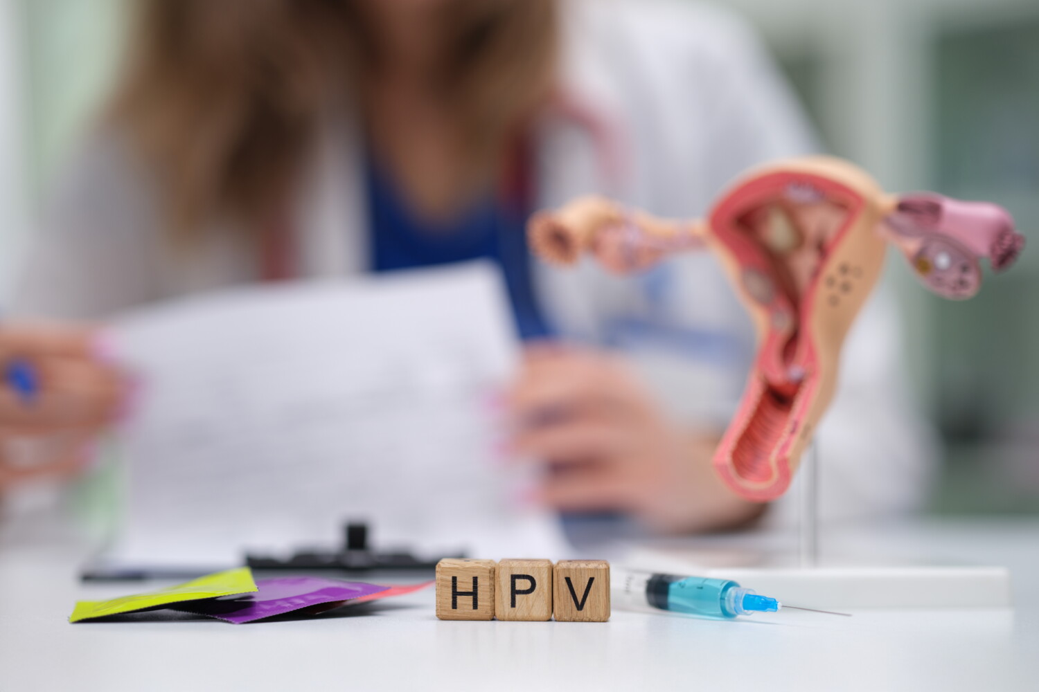 Napis HPV ułożony z drewnianych klocków, w tle model macicy, strzykawka i lekarka w białym fartuchu